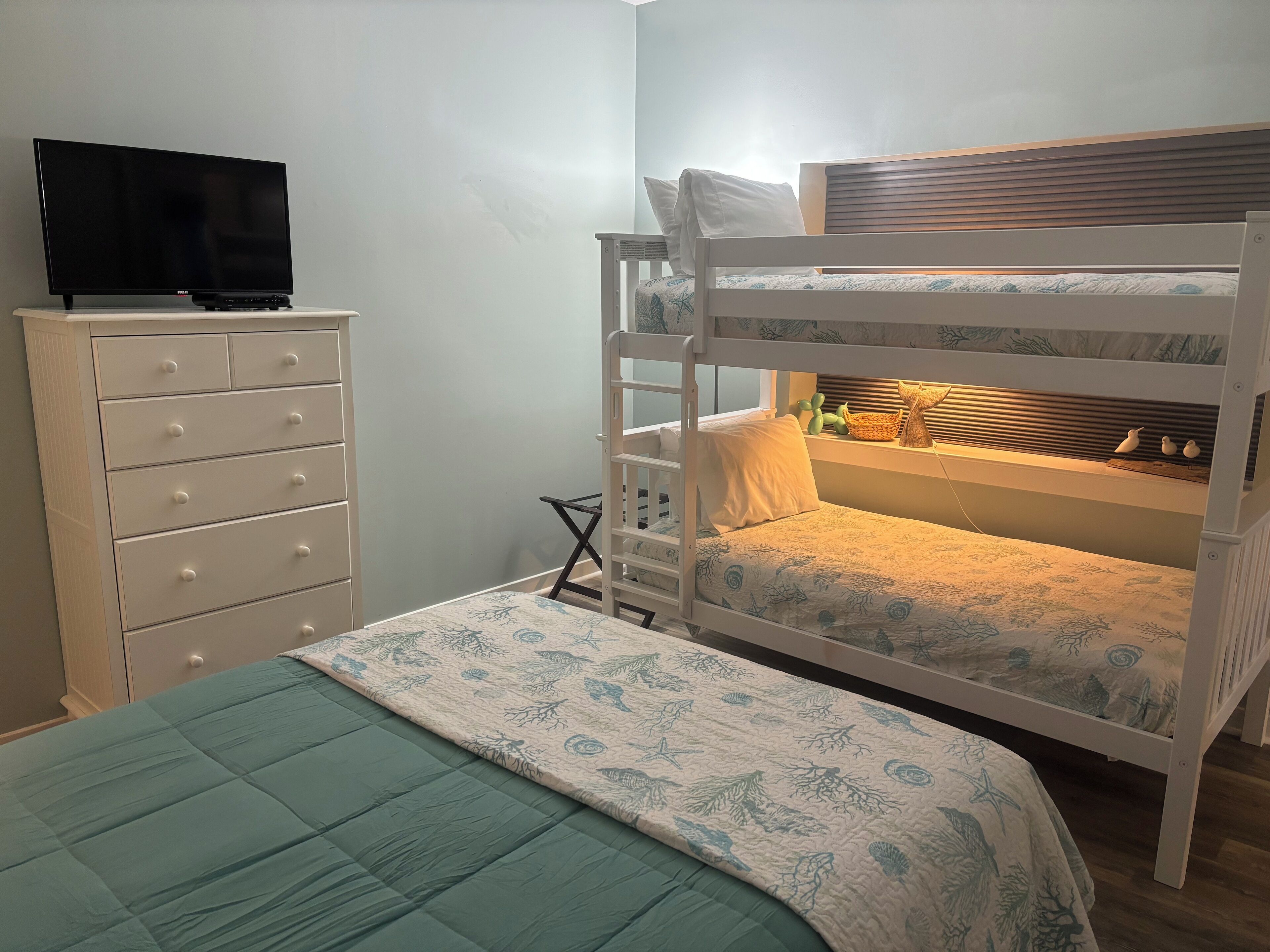A bunk bed with a lower bunk and a single bed, a dresser with drawers, a television on a stand, and a window with blinds.