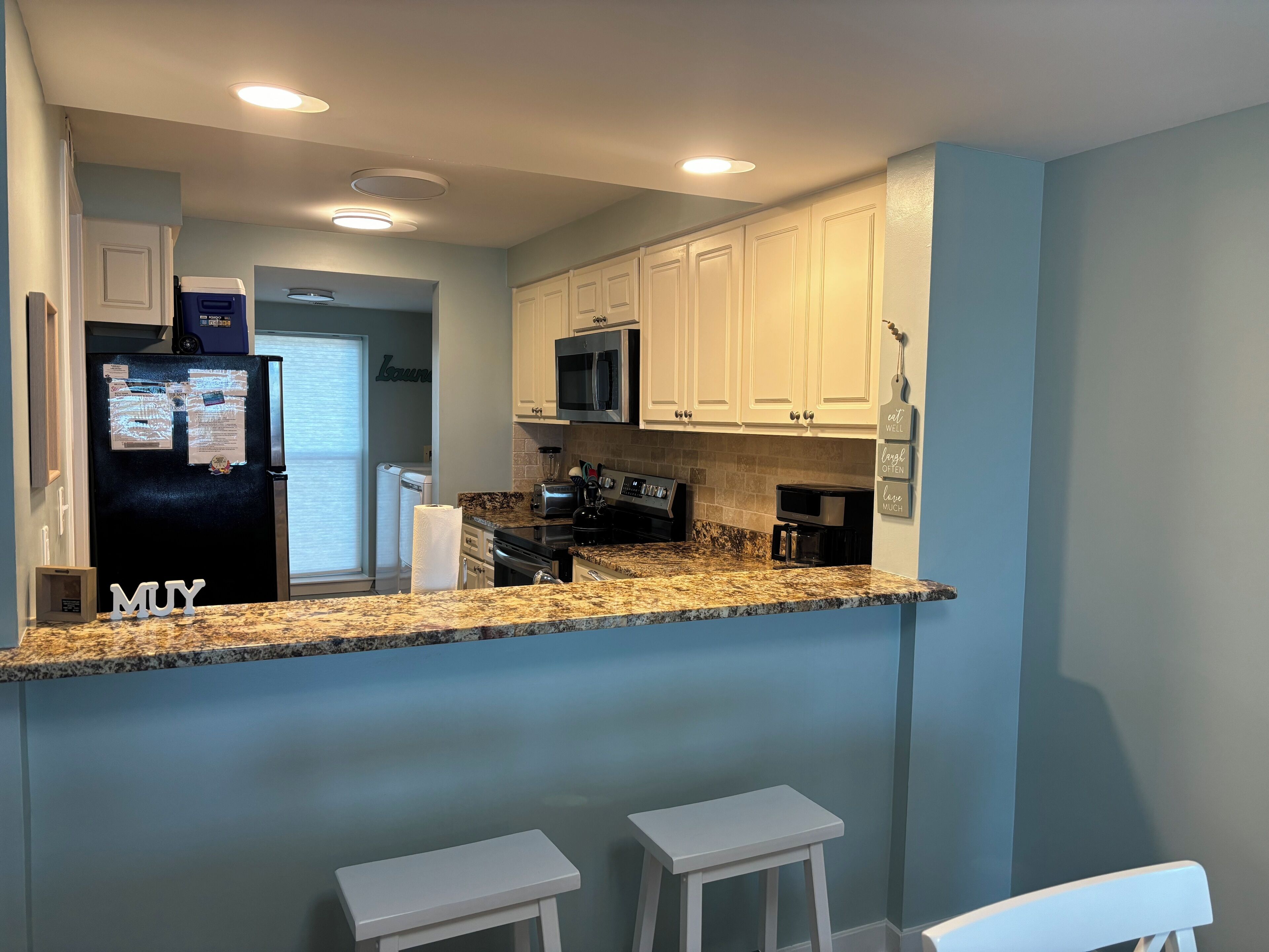 A kitchen with white cabinets, a black refrigerator, a microwave, and a granite countertop. There are two white stools and a window with blinds.