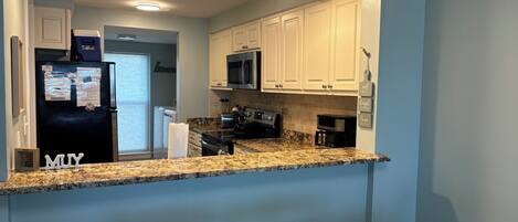 A kitchen with white cabinets, a black refrigerator, a microwave, and a granite countertop. There are two white stools and a window with blinds.