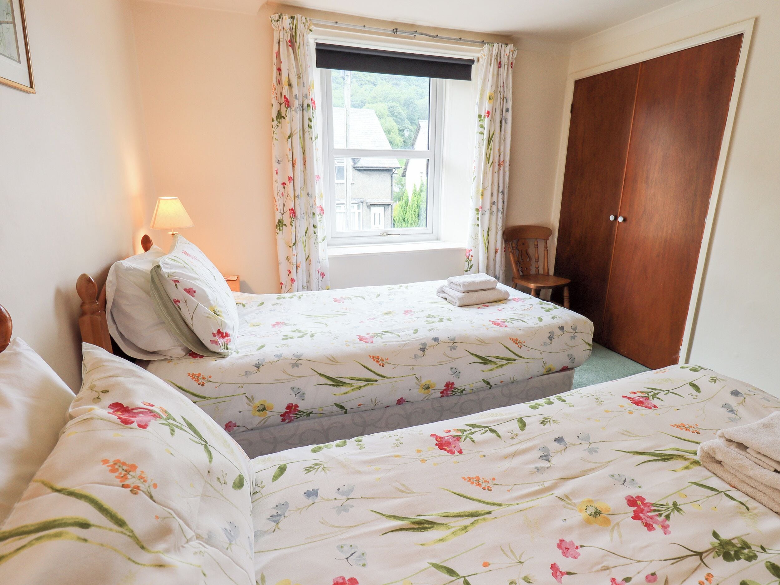 A bedroom with two beds, a window with floral curtains, a wooden chair, and a wooden wardrobe.