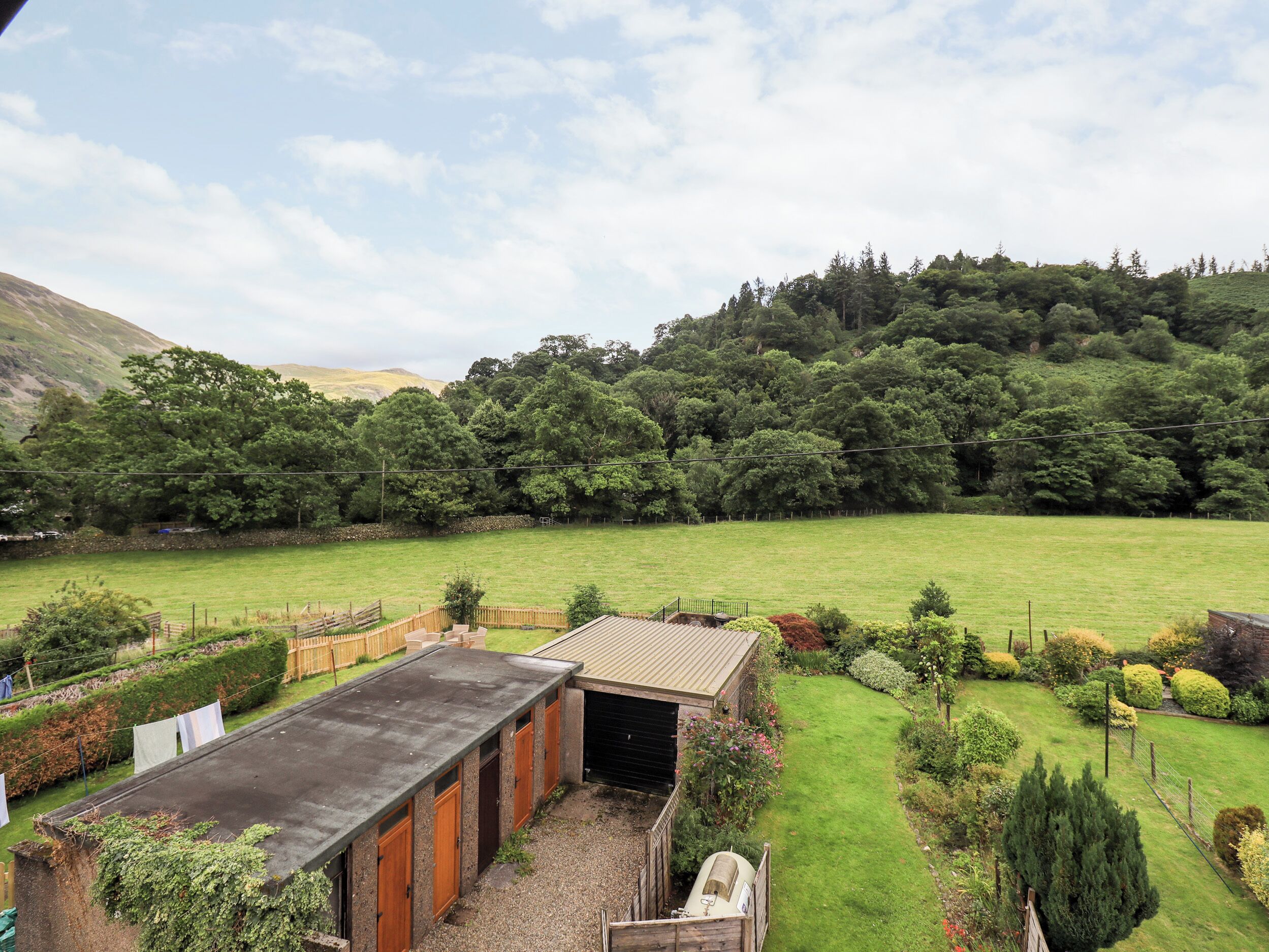 A rural property with a large open field, a wooden building with a sloped roof, and a well-maintained garden with various shrubs and trees.