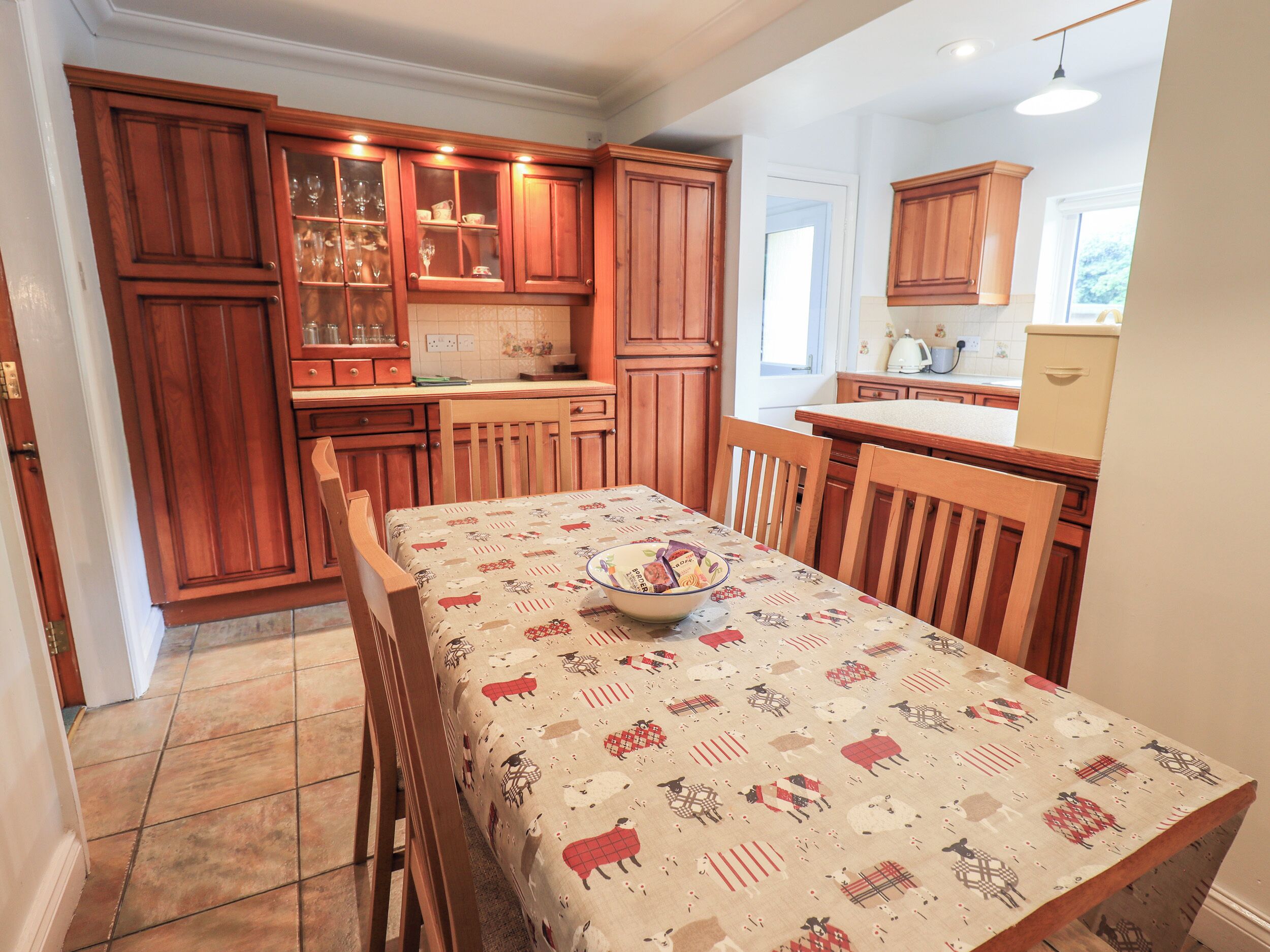 Une cuisine avec des meubles de rangement en bois, un sol carrelé et une table recouverte d’une nappe à motifs.