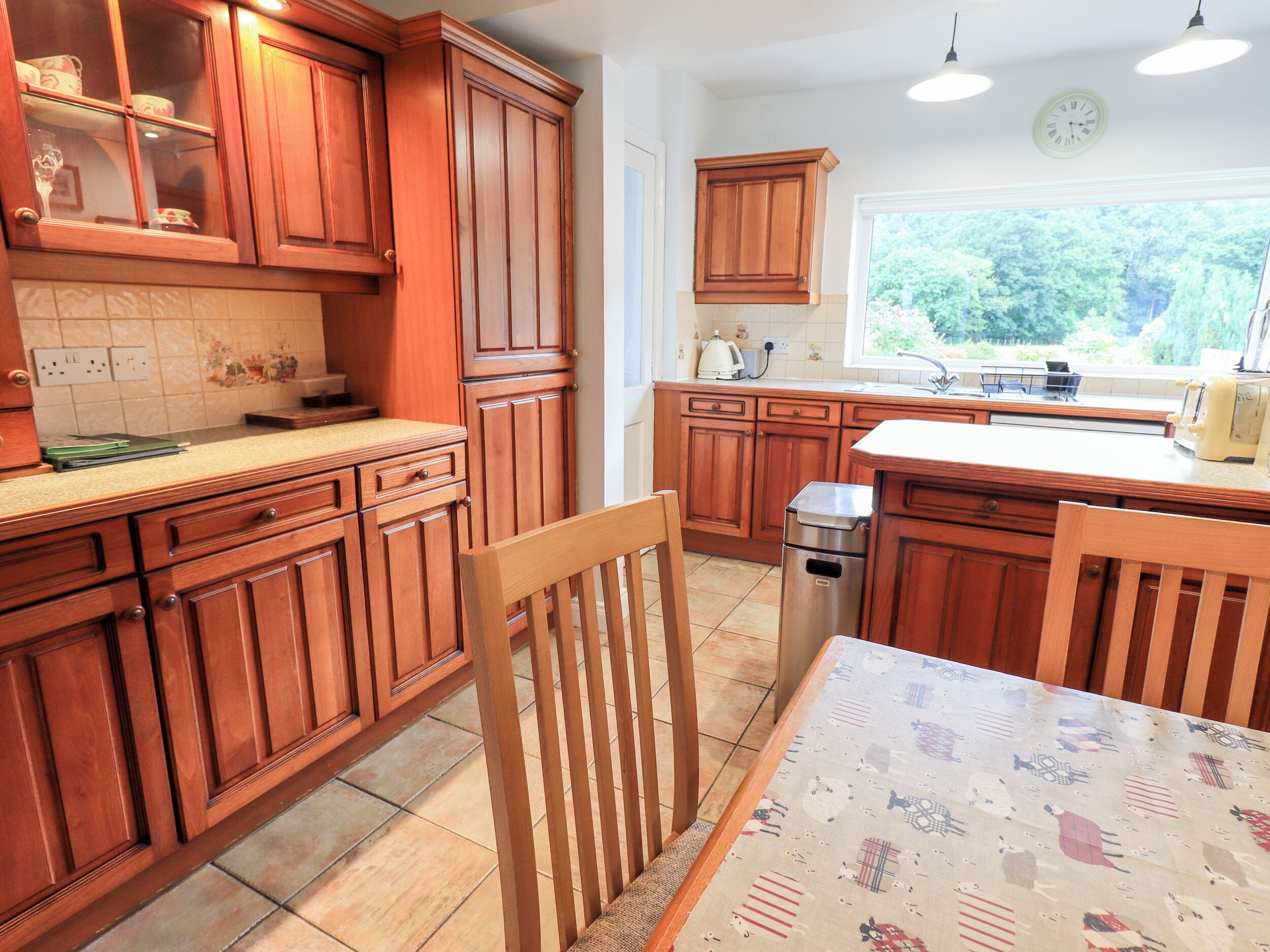 A kitchen with wooden cabinets, a stainless steel trash bin, a window with a view of greenery, and a table with a patterned tablecloth.