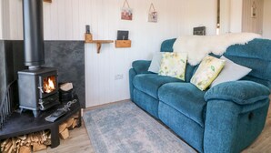A cozy living room with a wood-burning stove, a blue sofa with floral cushions, and a rustic wooden shelf against a white paneled wall. - Langdale Lodge (Okehampton)