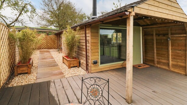 A wooden cabin with a glass door, a chimney, and a deck with a metal chair. - Langdale Lodge (Okehampton)
