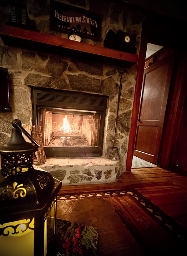 A stone fireplace with a lit fire, a clock above it, and a wooden door to the right.