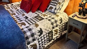Une chambre à coucher aux murs lambrissés, avec une courtepointe à carreaux, des coussins rouges et une table de chevet sur laquelle se trouvent une lampe et une petite décoration en forme d’arbre.