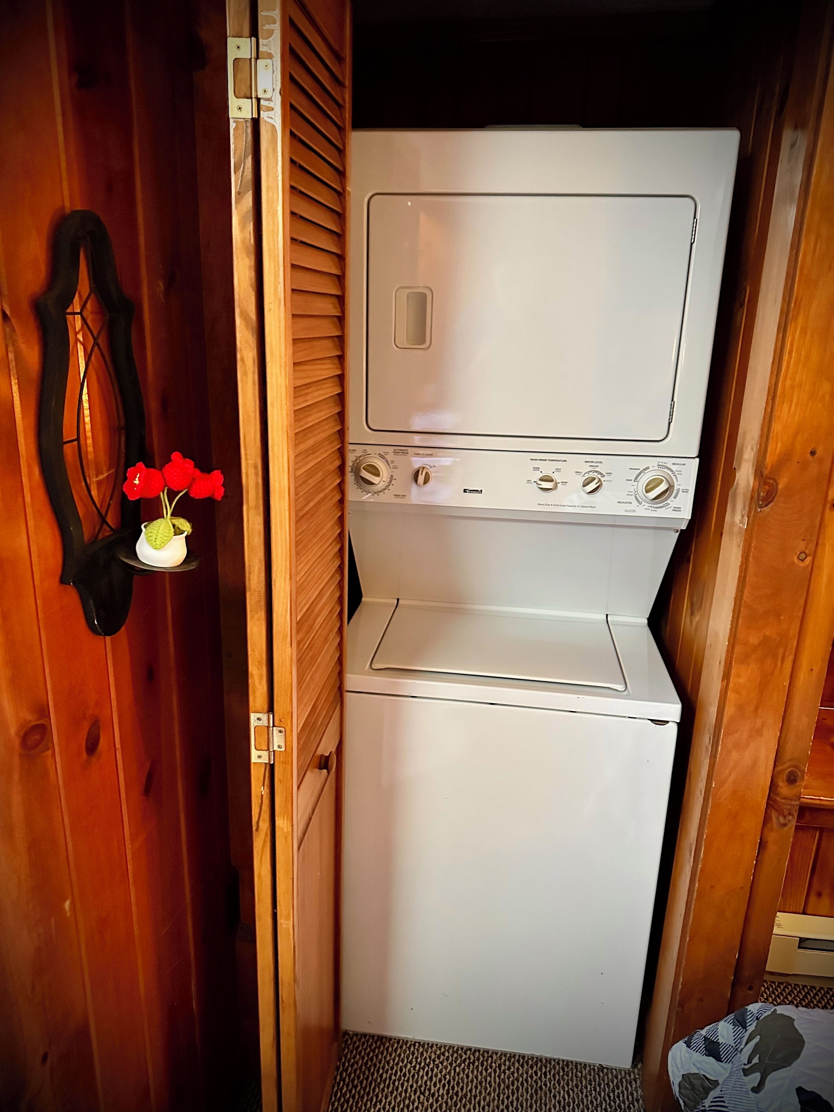 A compact laundry room with a white washer and dryer, wooden walls, and a small shelf with red flowers.