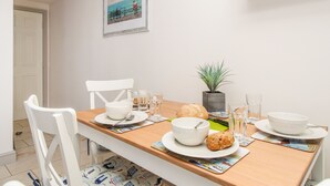 A dining area with a wooden table set for two, white chairs, a potted plant, and a framed picture on the wall. - Bliss On The Bay (Weymouth)