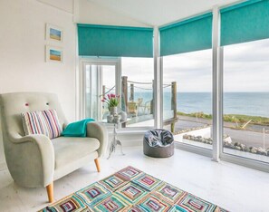A room with a large window overlooking the sea, a grey armchair with a colorful cushion, a patterned rug, and a small table with a vase of flowers. - The View (Port Isaac)