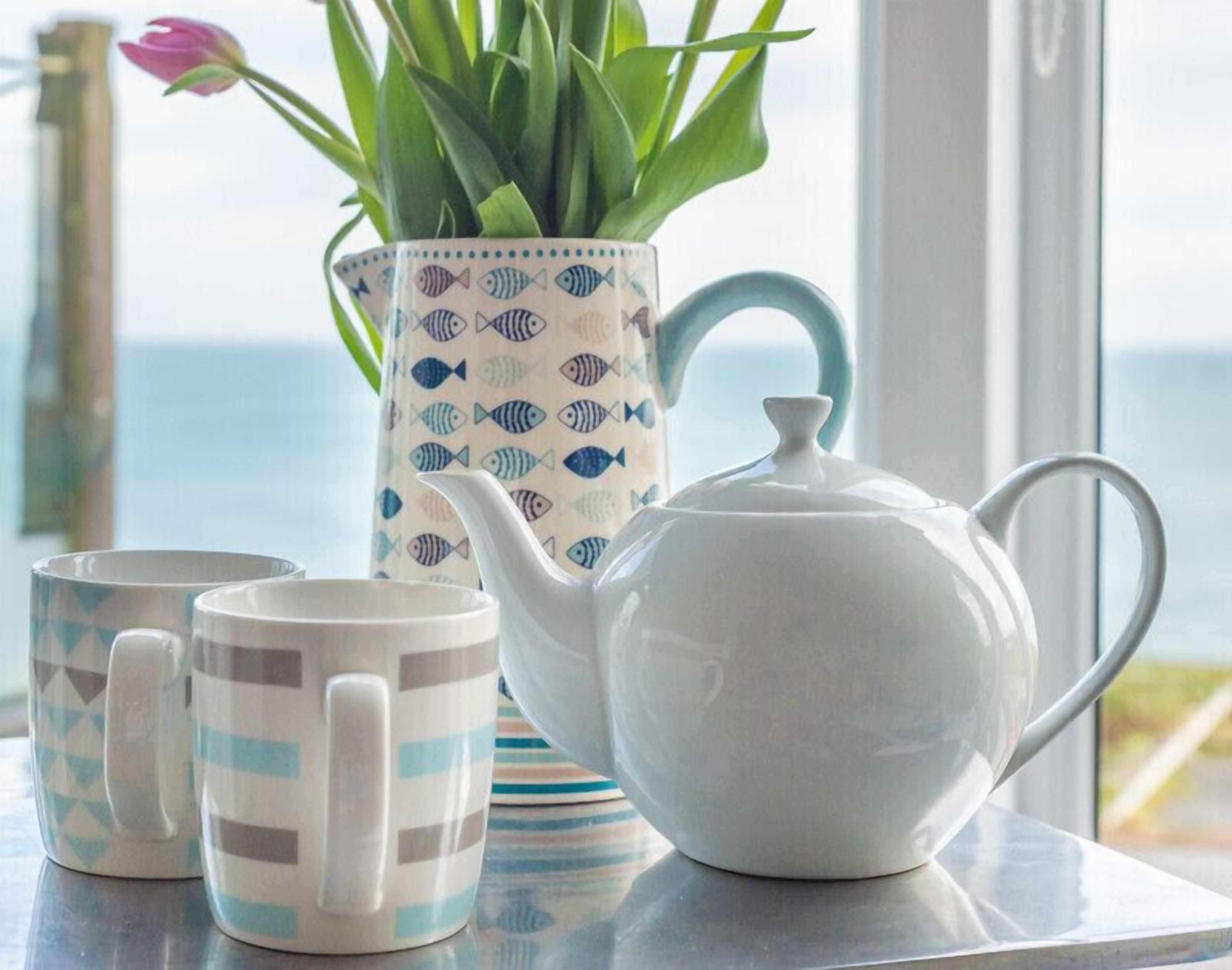A table with a white teapot, two striped mugs, and a fish-patterned vase with green plants.