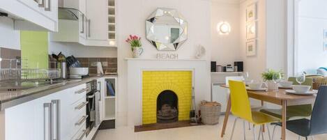 A modern kitchen with white cabinetry, a yellow brick fireplace, and a dining area with a wooden table and yellow chairs.