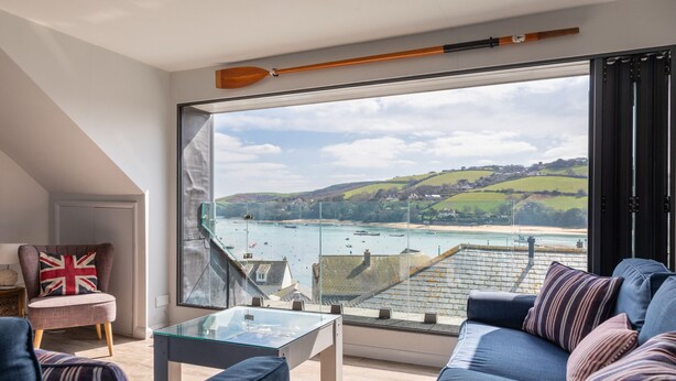 A modern living room with a glass coffee table, a sofa, and a chair with a British flag cushion. A large window offers a view of a coastal landscape with boats and buildings. - 2 Church Hill House (Salcombe)