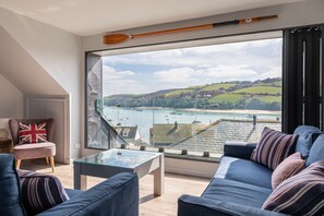 A modern living room with a glass coffee table, a sofa, and a chair with a British flag cushion. A large window offers a view of a coastal landscape with boats and buildings. - 2 Church Hill House (Salcombe)