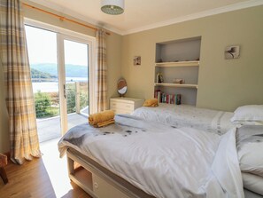 A bedroom with two beds, a window with curtains, a bedside table with a mirror, and a shelf with books.