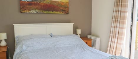 Een slaapkamer met een bed, nachtkastje, lamp, radiator en een schilderij van een kustlandschap.