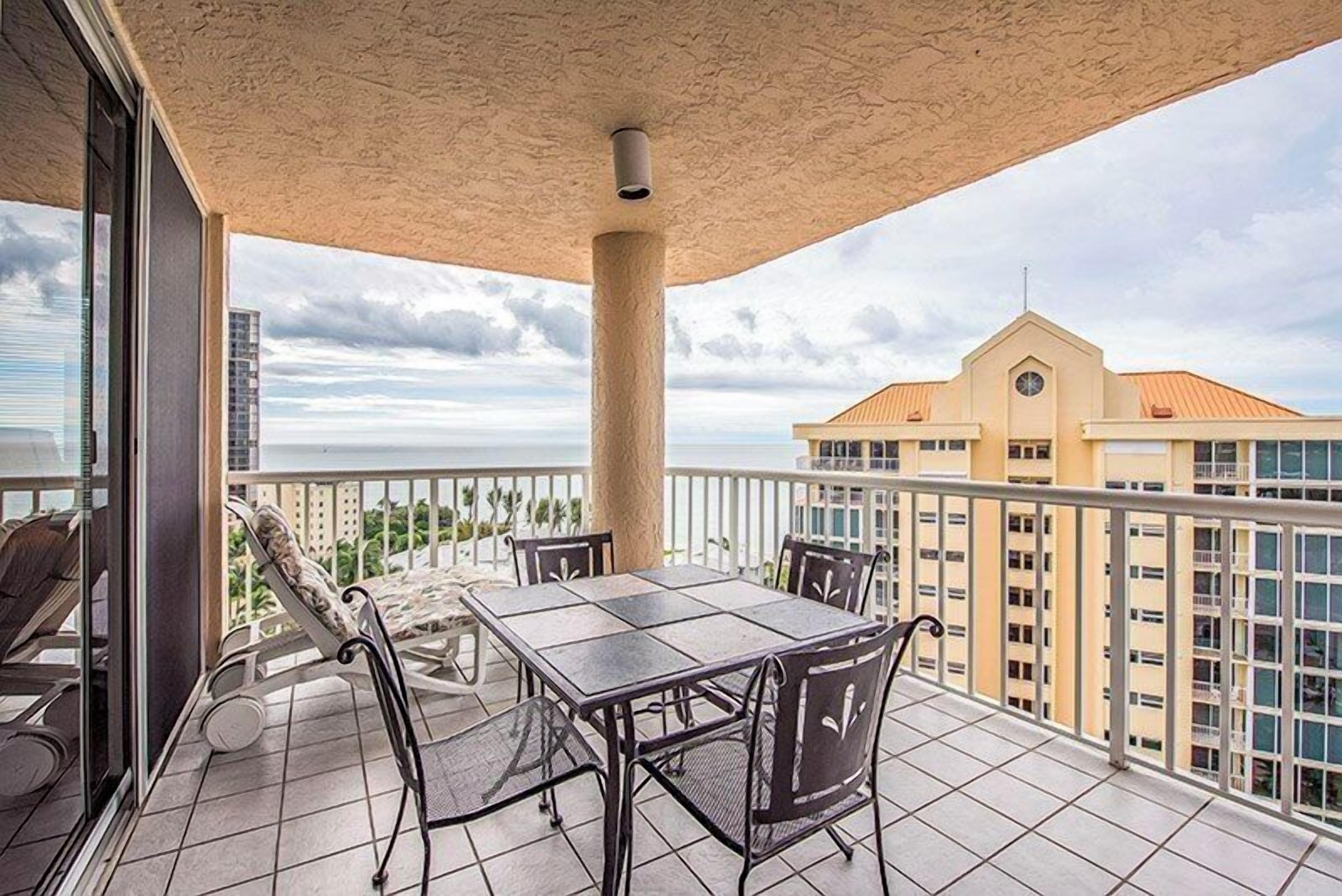 A balcony with a table and chairs, overlooking a cityscape and the ocean.