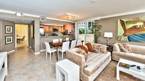 A spacious living room with a sofa, coffee table, and dining area. A kitchen is visible in the background. - Steps to the beach w/ a beautiful pool view CL0101 (Naples)