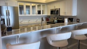 A modern kitchen with white cabinetry, stainless steel appliances, a marble countertop, and bar stools. - Superb Gulf views in a renovated unit - CL0302 (Naples)