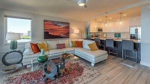 Una sala moderna con un sofá modular, una mesa de centro y un cuadro grande. Hay una zona de cocina con taburetes de bar y lámparas colgantes.