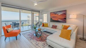 Una sala de estar moderna con un sofá blanco, un sillón naranja y una mesa de centro de vidrio. Hay un ventanal con vistas a edificios y un balcón.