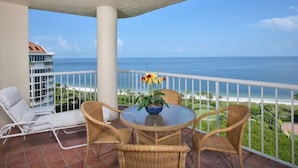 A balcony with a view of the ocean, a white chair, a wicker table with a vase of flowers, and a white railing. - Delightful Gulf views from the 11th floor!-WS1102 (Naples)