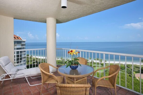 A balcony with a view of the ocean, a white chair, a wicker table with a vase of flowers, and a white railing.