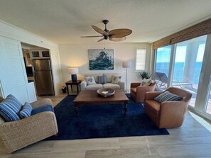 A spacious living room with a ceiling fan, a large painting, and a view of the ocean through the windows.