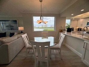 A modern kitchen with a dining area, featuring a round glass table and white chairs, a large window with a city view, and a ceiling light fixture. - Views of palm trees, sand and the Gulf! - WS0304 (Naples)