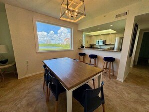 Un coin repas avec une table en bois et des chaises noires, une grande fenêtre donnant sur les nuages et les bâtiments, et une cuisine en arrière-plan.