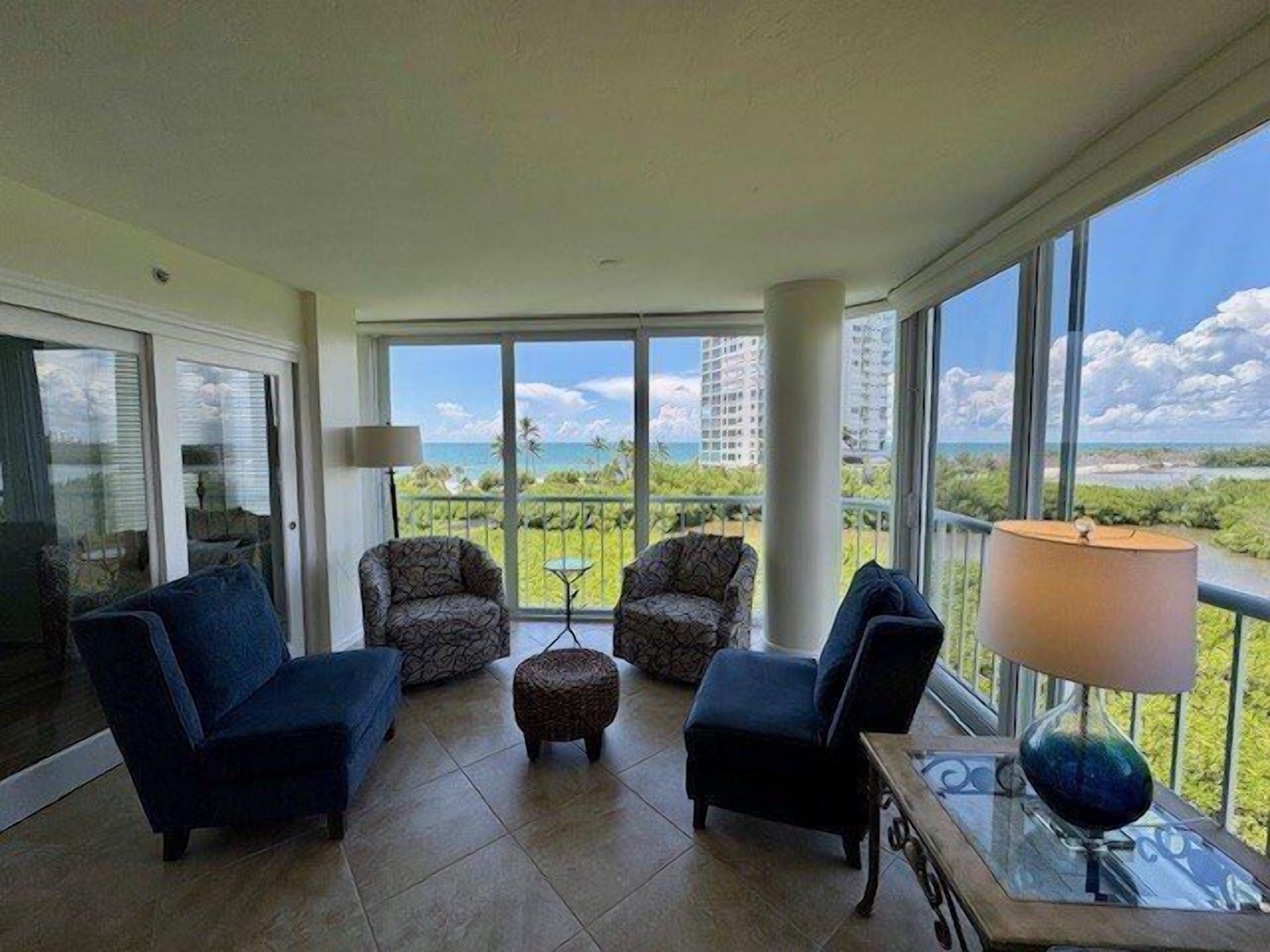 A spacious balcony with a view of the ocean and buildings, furnished with two armchairs, a small table, and a lamp.