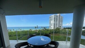A covered outdoor area with a round table and four chairs, overlooking a beach and a body of water with a tall building in the background. - Gulf of Mexico view from your living room-WS0503 (Naples)