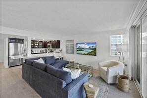 Una sala moderna con un sofá gris, una mesa de centro de vidrio y un sillón blanco. Se ve una cocina al fondo, con armarios oscuros y un ventanal grande.