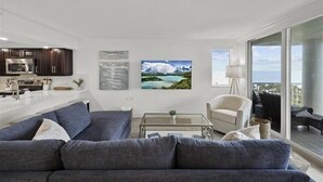 Un soggiorno moderno con un divano grigio, un tavolino da caffè e una TV. È presente un angolo cottura con pensili scuri e un forno a microonde. Al muro, un quadro raffigura un paesaggio montano.