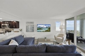 Una sala de estar moderna con un sofá gris, una mesa de centro y un televisor. Hay una zona de cocina con armarios oscuros y un microondas. En la pared, un cuadro de un paisaje montañoso.