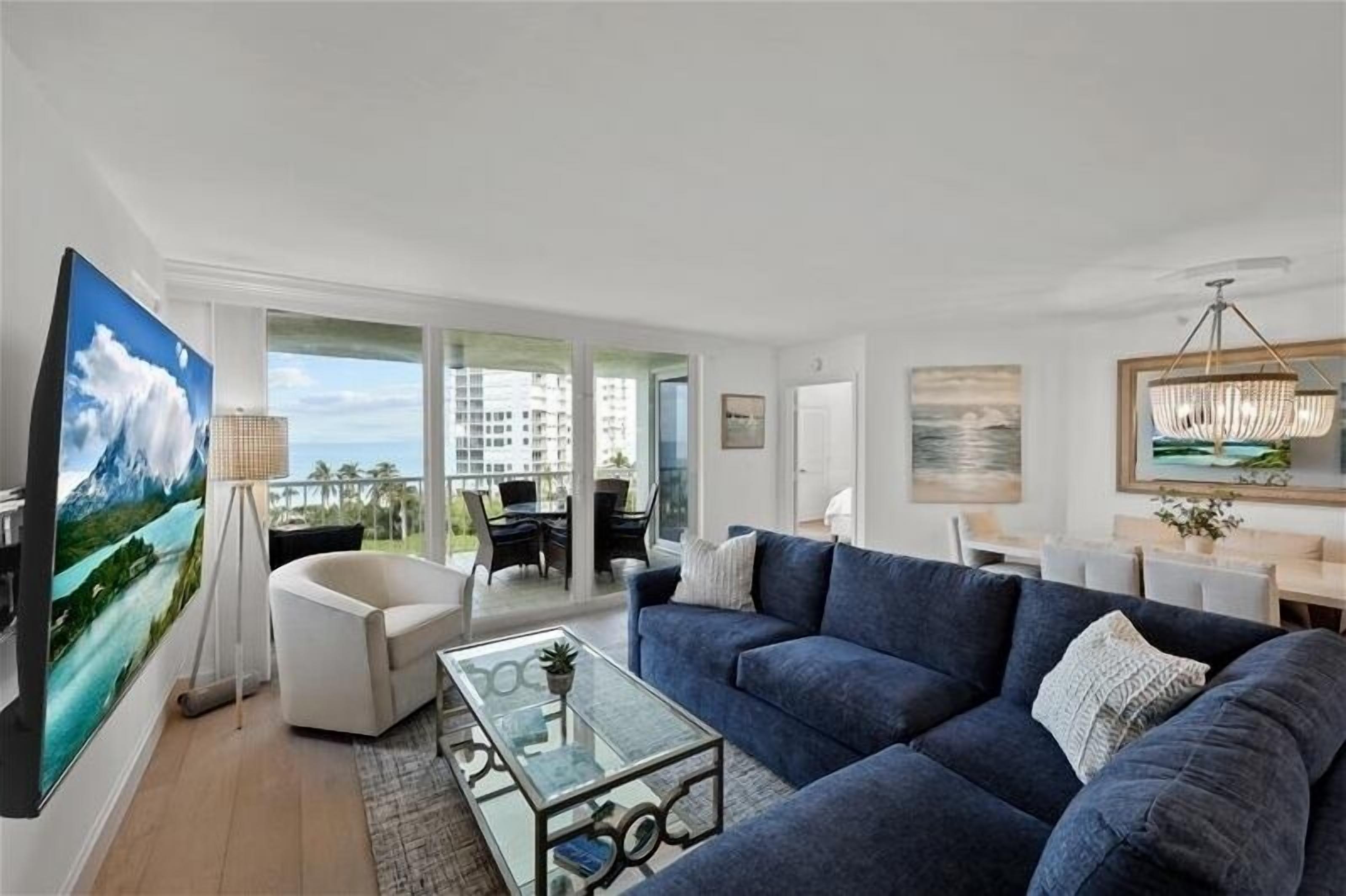 Ein modernes Wohnzimmer mit einem großen Flachbildfernseher, einem blauen Sofa, einem Glastisch und einem Balkon mit Gartenmöbeln.