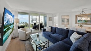 Un soggiorno moderno con una grande TV a schermo piatto, un divano blu, un tavolino in vetro e un balcone con mobili da esterno.