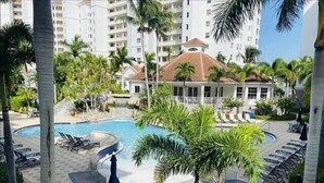 Zona de piscina en un resort con sillones, palmeras y un edificio al fondo.