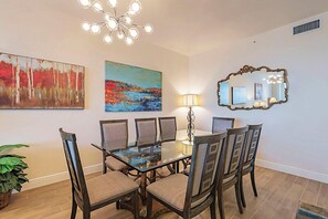 A dining area with a glass table surrounded by chairs, a chandelier, and two paintings on the wall. - 2 blocks from the beach with bay and gulf views - Reg 2-1004 (Naples)