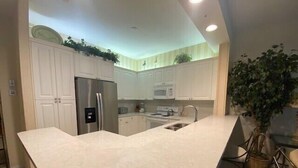 A modern kitchen with white cabinetry, a stainless steel refrigerator, a microwave, and a sink. There is a large island with a chair and a potted plant. - Great location! 2 blocks from the beach with beautiful pool views -Reg 5-404 (Naples)