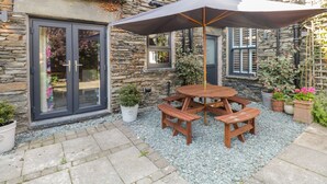 Ein Steinhaus mit Terrasse, das einen Holztisch und Bänke sowie üppige Pflanzkübel beherbergt.