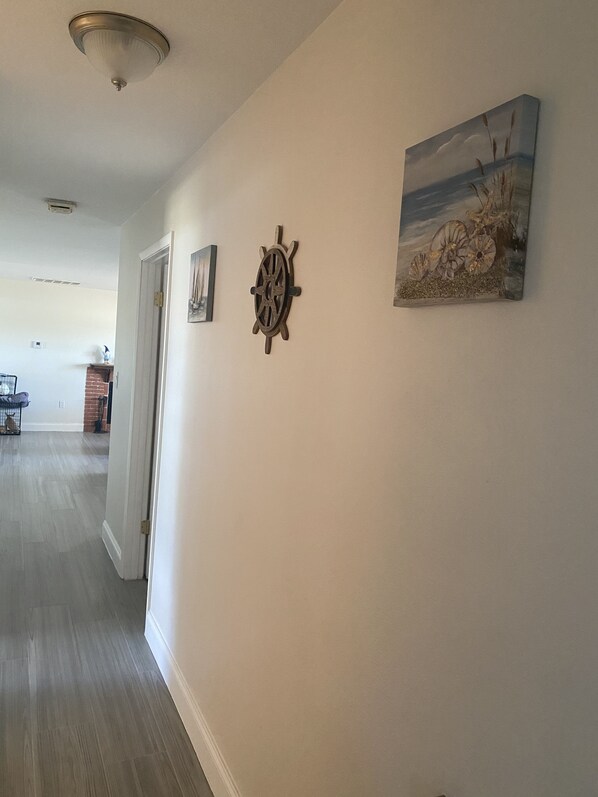 Un couloir avec un plancher en bois, une porte, un gouvernail de bateau fixé au mur et un tableau sur le thème de la plage.