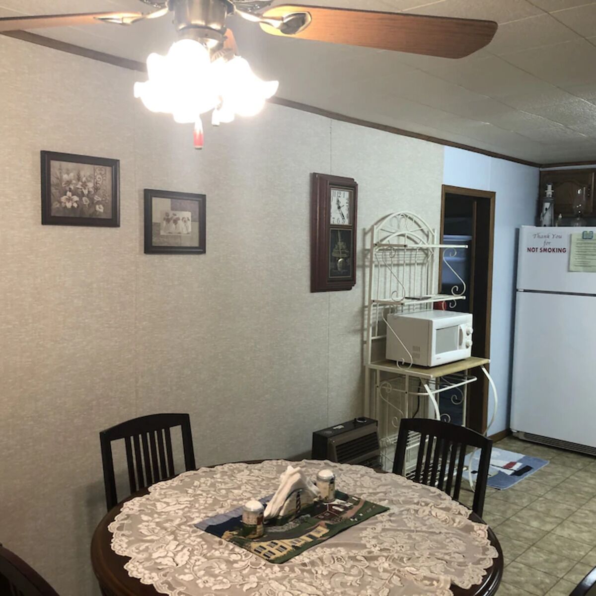 A dining area with a round table covered by a lace tablecloth, two chairs, a ceiling fan, and a clock on the wall.