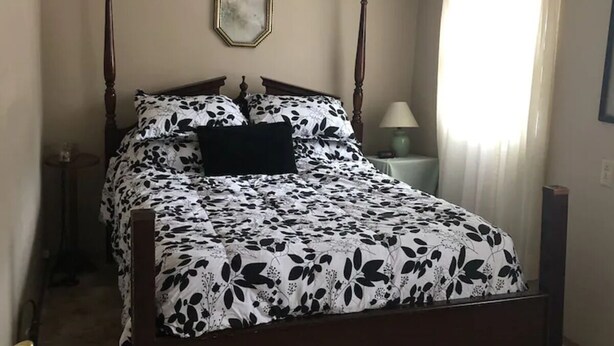 A bedroom with a four-poster bed, a nightstand, a lamp, and a window with curtains.