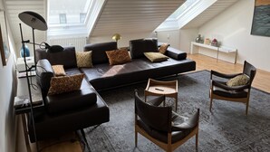 A modern loft apartment with a large black leather sofa, two leather chairs, a standing lamp, and a skylight.