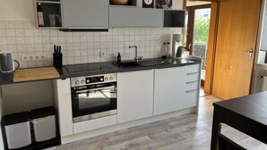 A modern kitchen with white cabinets, a black countertop, and a built-in oven. There are two trash bins and a wooden cutting board on the counter.