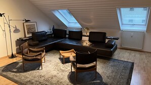 A modern loft-style living room with a sectional sofa, two armchairs, a floor lamp, and a large window with skylights.