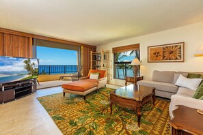 A spacious living room with a large TV, comfortable seating, a wooden coffee table, and a view of the ocean.