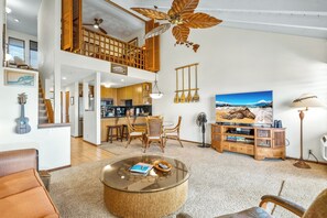 Een ruime woonkamer met een grote ronde salontafel, houten meubels, een plafondventilator en een grote televisie aan de muur.