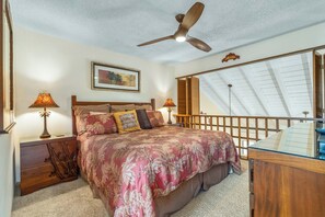 A bedroom with a bed, two bedside lamps, a ceiling fan, a framed picture, and a view of the outside through a window.